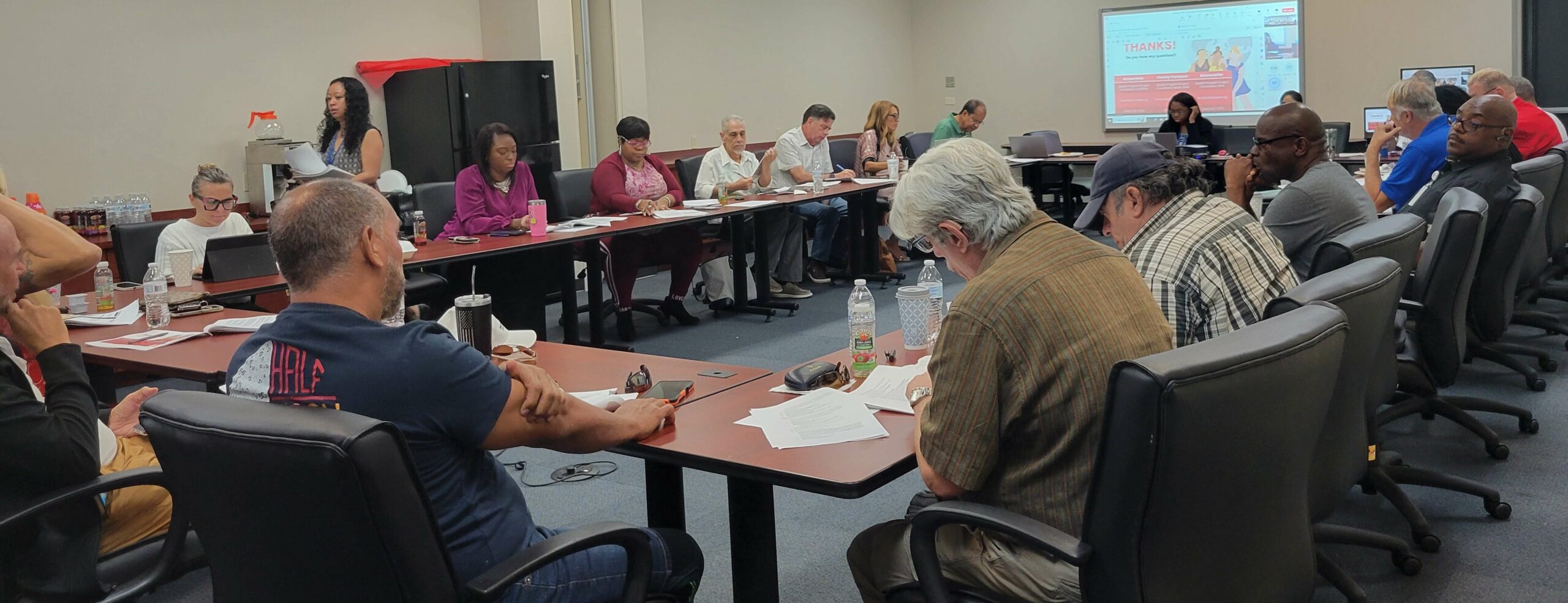 A group of people are gathered in a conference room, seated around a large U-shaped table. Some individuals have laptops or notebooks in front of them, while others appear to be listening attentively. A woman is standing, possibly presenting information, and there is a screen at the front of the room displaying a presentation slide that includes the text "THANKS!" This indicates that the session might be concluding or there has been an opportunity for questions. The environment appears collaborative and focused on discussion or training.