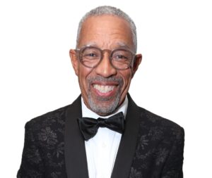 A smiling older man with graying close-cropped hair and a beard is seen in the image. He is wearing round eyeglasses and is dressed in a formal black tuxedo jacket adorned with a subtle floral pattern, a white dress shirt, and a black bow tie.