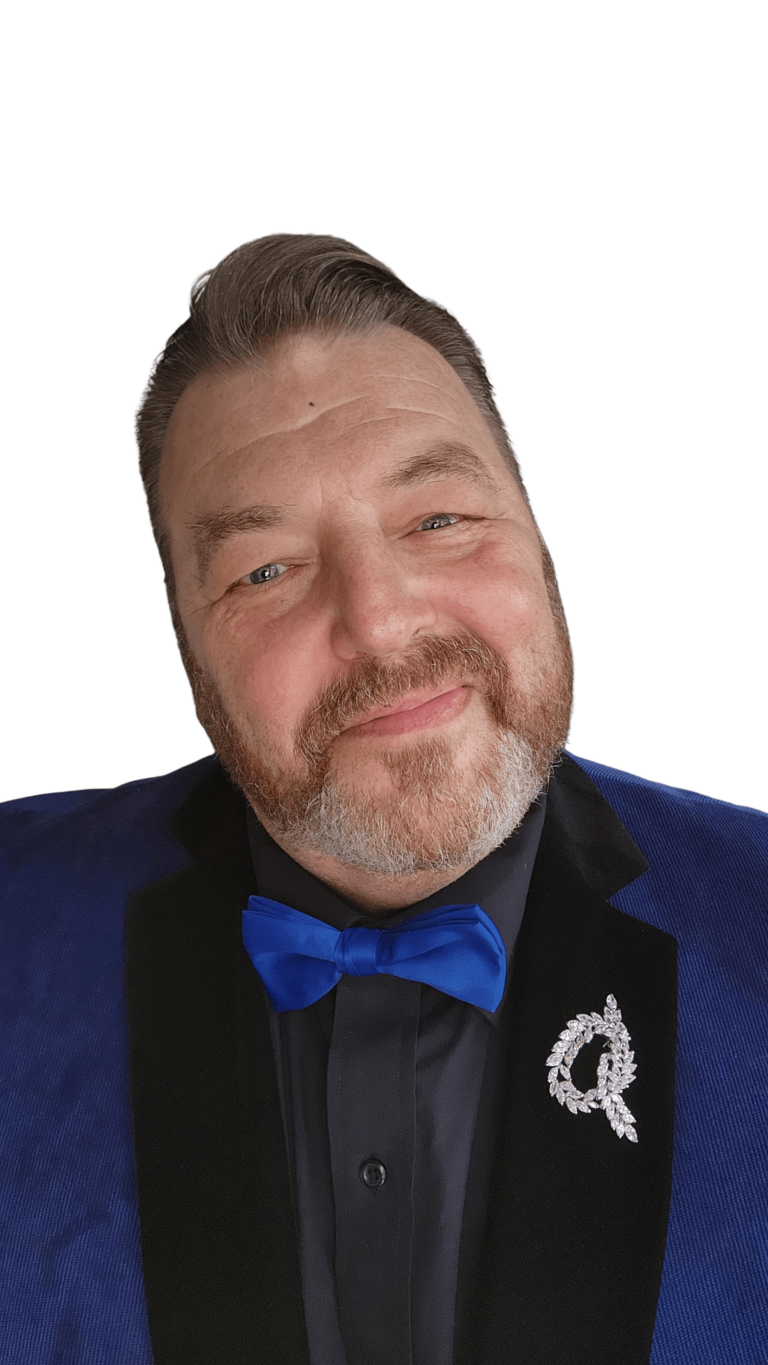 A man with a neatly groomed beard and mustache is smiling at the camera. He is wearing a blue suit jacket, a black shirt, and a blue bow tie. There is a decorative brooch pinned to his lapel. The background is slightly blurred with what appears to be a modern indoor setting.