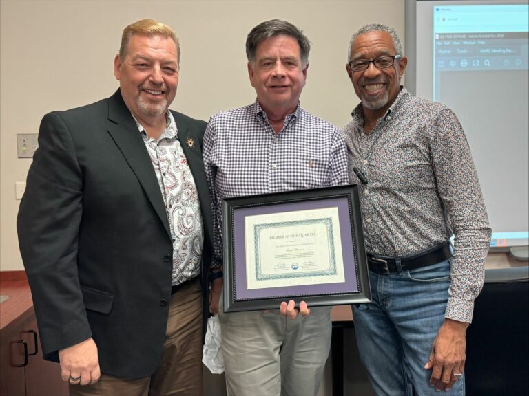 The image shows three individuals standing side by side. One of them is holding a framed certificate titled "Member of the Quarter." The individuals are dressed in business casual attire, with the person on the left wearing a blazer and patterned shirt, the person in the middle wearing a checkered shirt, and the person on the right wearing a patterned button-up shirt. They appear to be in an office or meeting room setting, with a projection screen visible in the background.