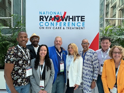 This image appears to be from a conference or event focused on HIV care and treatment. The backdrop shows the text "NATIONAL RYAN WHITE CONFERENCE ON HIV CARE & TREATMENT" in blue and red lettering. In front of this backdrop, there's a group of seven people posing together. They seem to be attendees or organizers of the conference. The group is diverse in terms of age, gender, and ethnicity. They're all smiling and appear to be in a positive, friendly mood. The setting looks like it could be in a conference center or hotel lobby, with some greenery visible in the background behind the conference banner. This image shows a group of people at the "National Ryan White Conference on HIV Care & Treatment." The conference is a key event for HIV/AIDS professionals, focusing on advancements in care and fostering collaboration among healthcare providers, researchers, and policymakers. It's named after Ryan White, a national symbol of HIV/AIDS awareness. The event emphasizes improving care quality and discussing relevant healthcare policies.