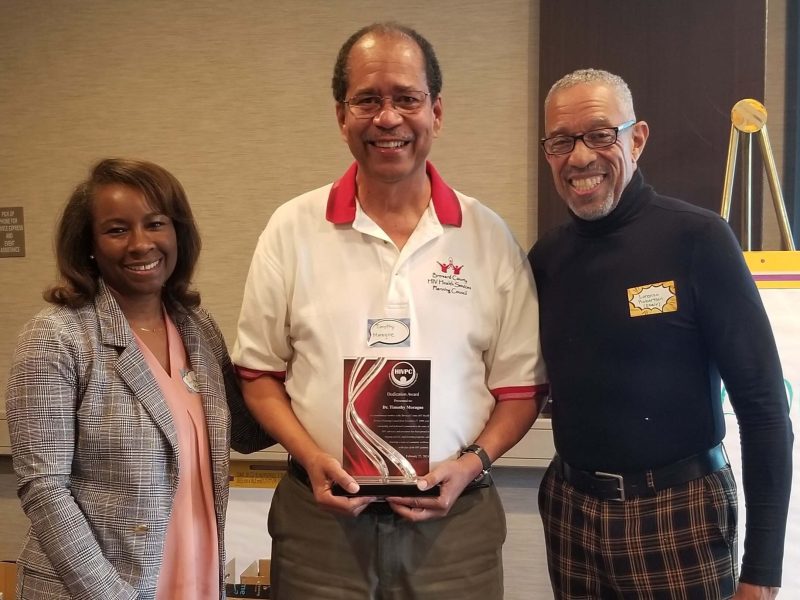 Three people standing together smiling; the person in the middle is holding an award. The woman on the left is wearing a blazer, light-colored blouse, and dark pants. The man in the center is wearing a white polo shirt with red trim, khaki pants, and is holding an award plaque. The man on the right is wearing a black turtleneck and plaid pants.