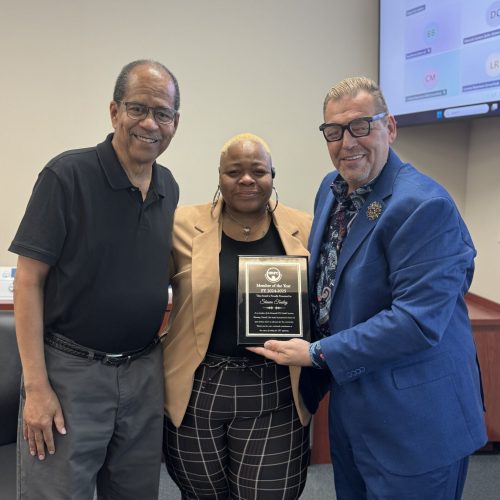Three council members stand in a conference room, smiling toward the camera. The central figure, a woman in a tan blazer and black shirt, holds a black and gold plaque reading “Member of the Year FY 2023–2024 Simone Finley.” A man in a blue suit and patterned shirt to her right presents the plaque, while a man in a black polo and gray pants stands to her left.