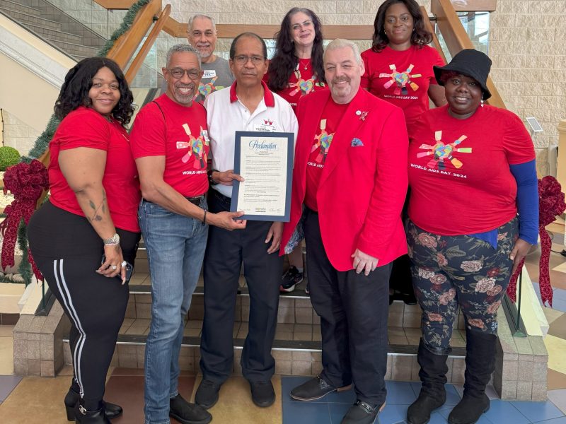 A group of nine members of the Broward County HIV Health Services Planning Council pose indoors on a staircase, most wearing matching red “World AIDS Day 2023” T-shirts. One person in a white polo holds a framed proclamation, and another in a red blazer stands beside them. They smile in front of holiday garland and colorful artwork, celebrating their shared mission